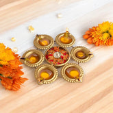 A set of two golden glossy metal diya urlis with a floral design, containing lit candles, displayed on a wooden surface with flowers around.